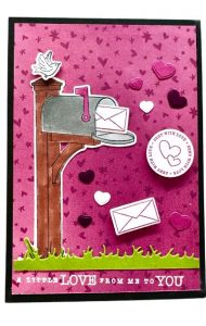 Handgefertigte Karte zum Valentinstag basteln mit einem 3D-Briefkastenmotiv, umgeben von ausgestanzten Herzen und einem Stempelaufdruck, der sagt 'A little love from me to you'. Der Hintergrund ist in einem lebhaften Pinkton mit Herzen verziert und am unteren Rand ist ein grüner Papierstreifen, der Gras darstellt